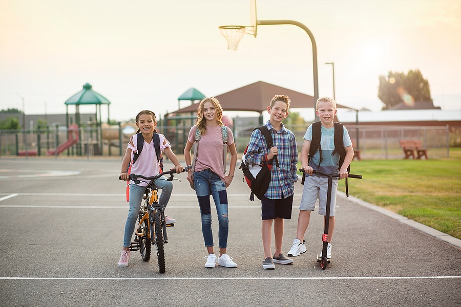 Group of elementary school kids in Norfolk smiling after school with backpacks, bike, and scooter on a playground.